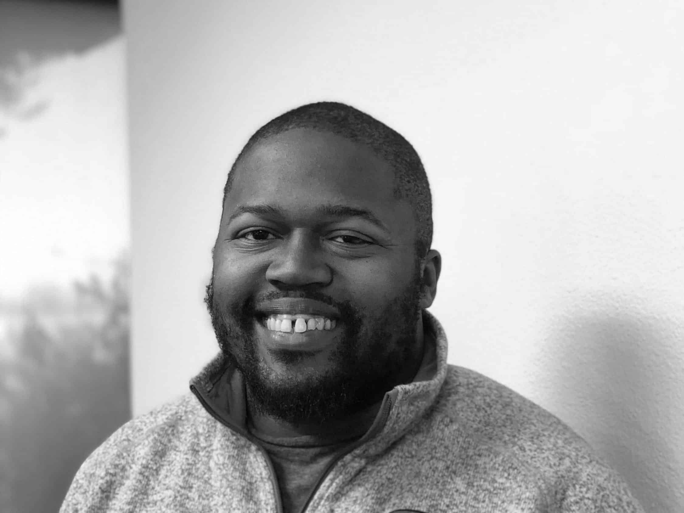Black and white portrait of a smiling man with a beard, wearing a hoodie, against a textured background.