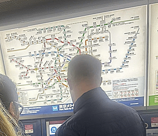 Two people stand in front of a large, illuminated subway map with various colored lines and station names. One person's back is visible as they look at the map, possibly planning their route.