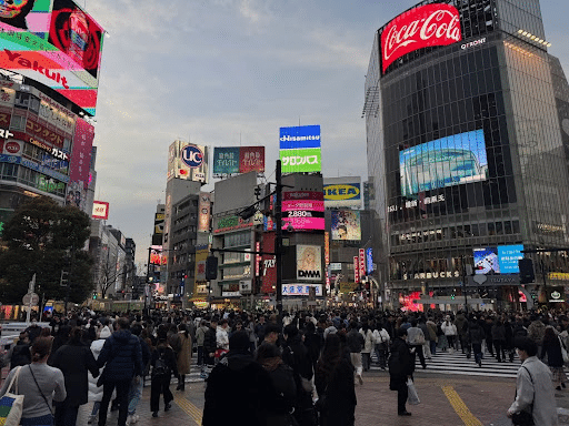A bustling urban scene at Shibuya Crossing in Tokyo, crowded with people walking in various directions. Surrounding buildings display large, colorful advertisements and digital billboards against a cloudy sky.