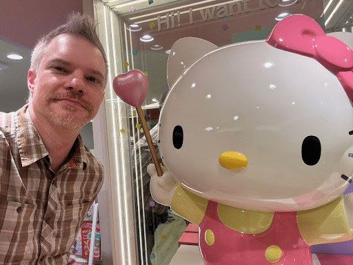 A man with short hair and a beard stands smiling next to a large statue of a character with a bow on its ear, holding a heart-shaped wand. The background features bright store lighting and colorful decorations.
